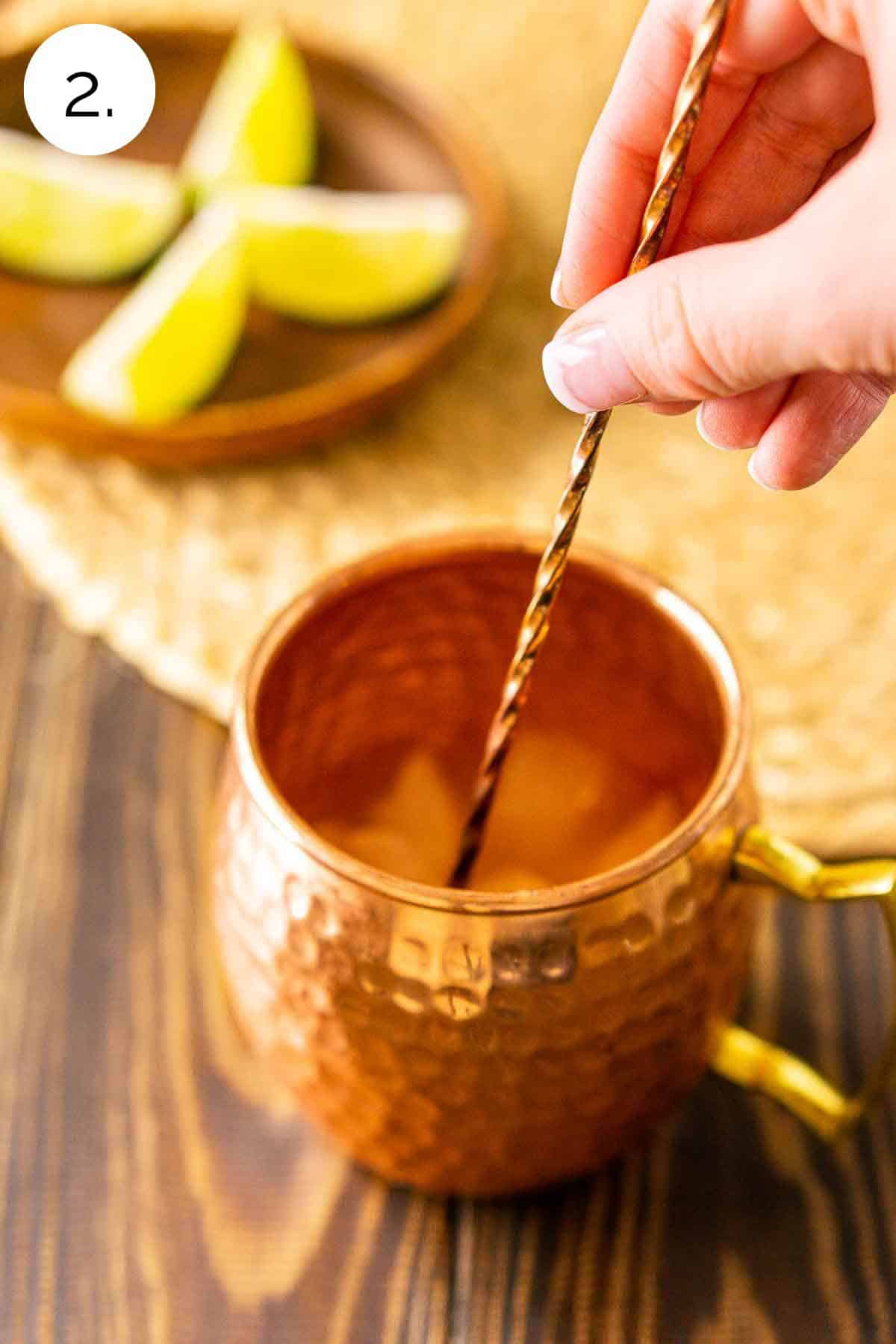 A hand stirring the lime juice, bourbon and ice in a copper mug with a bar spoon on a wooden surface.