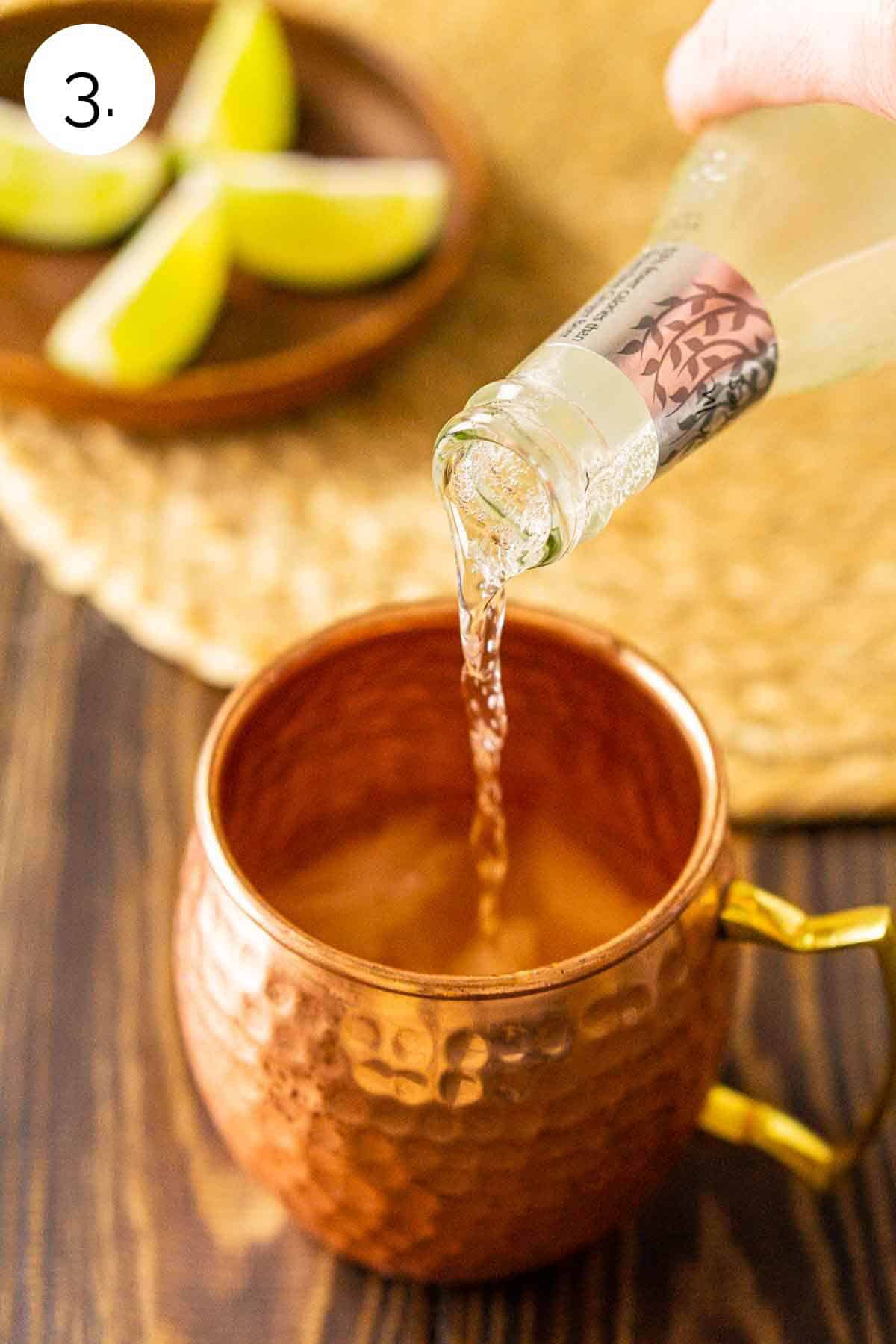 A hand pouring in the ginger beer into the copper mug with the ice and other ingredients on a wooden surface.