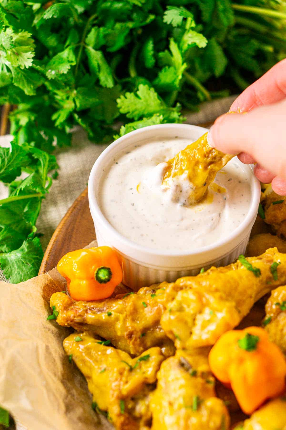 A hand dipping a mango-habanero wing into a white cup filled with ranch dressing and a bundle of cilantro behind it.