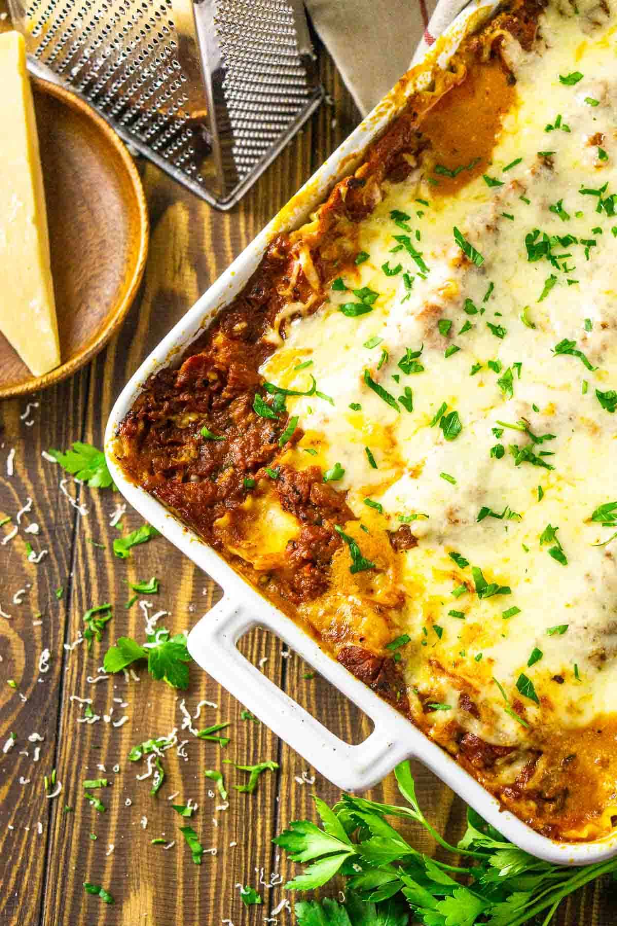 An aerial view of a baking dish filled with the meatball lasagna with Parmesan cheese and parsley scattered around it on a wooden surface.