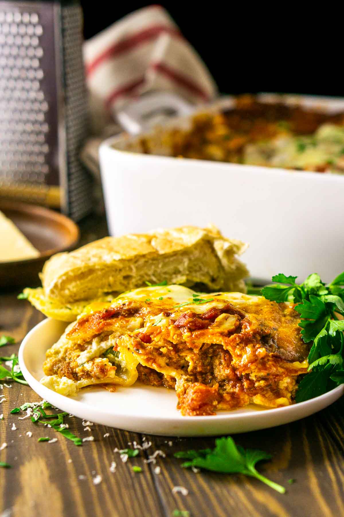 A slice of the meatball lasagna on a small white plate with grated Parmesan and chopped parsley in front.