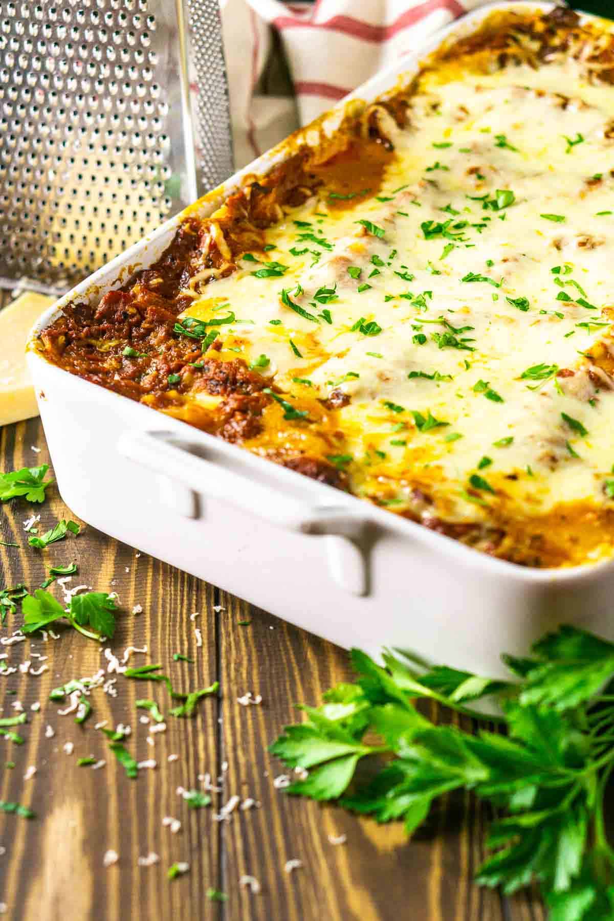 A pan of the meatball lasagna on a brown wooden surface with a cheese grater and wedge of Parmesan in the background.