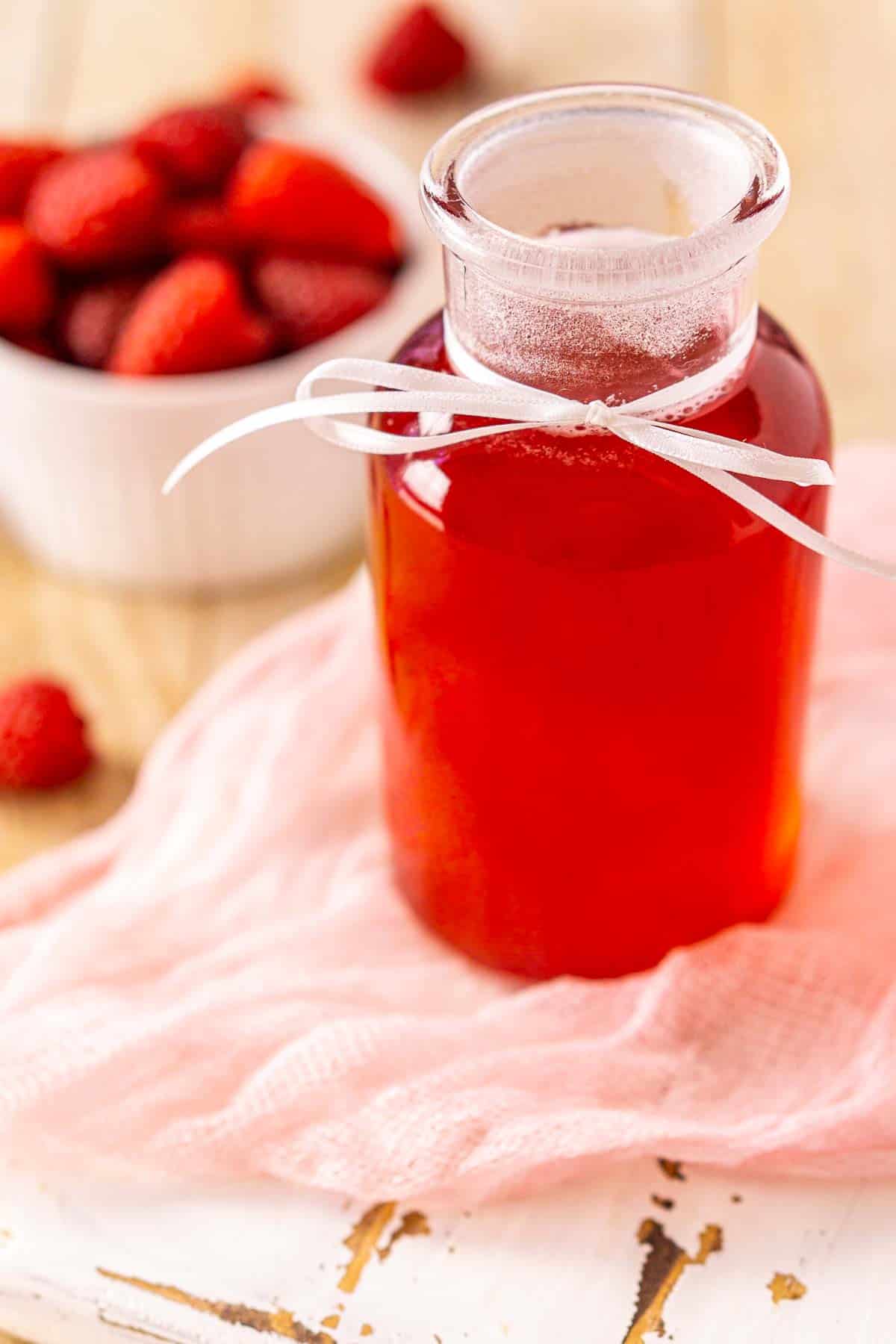 A close-up of the raspberry simple syrup in a glass jar on a white wooden tray with a piece of pink clothe.