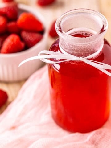 A bottle of raspberry simple syrup on a white tray with a piece of pink clothe and a small bowl of berries to the left.