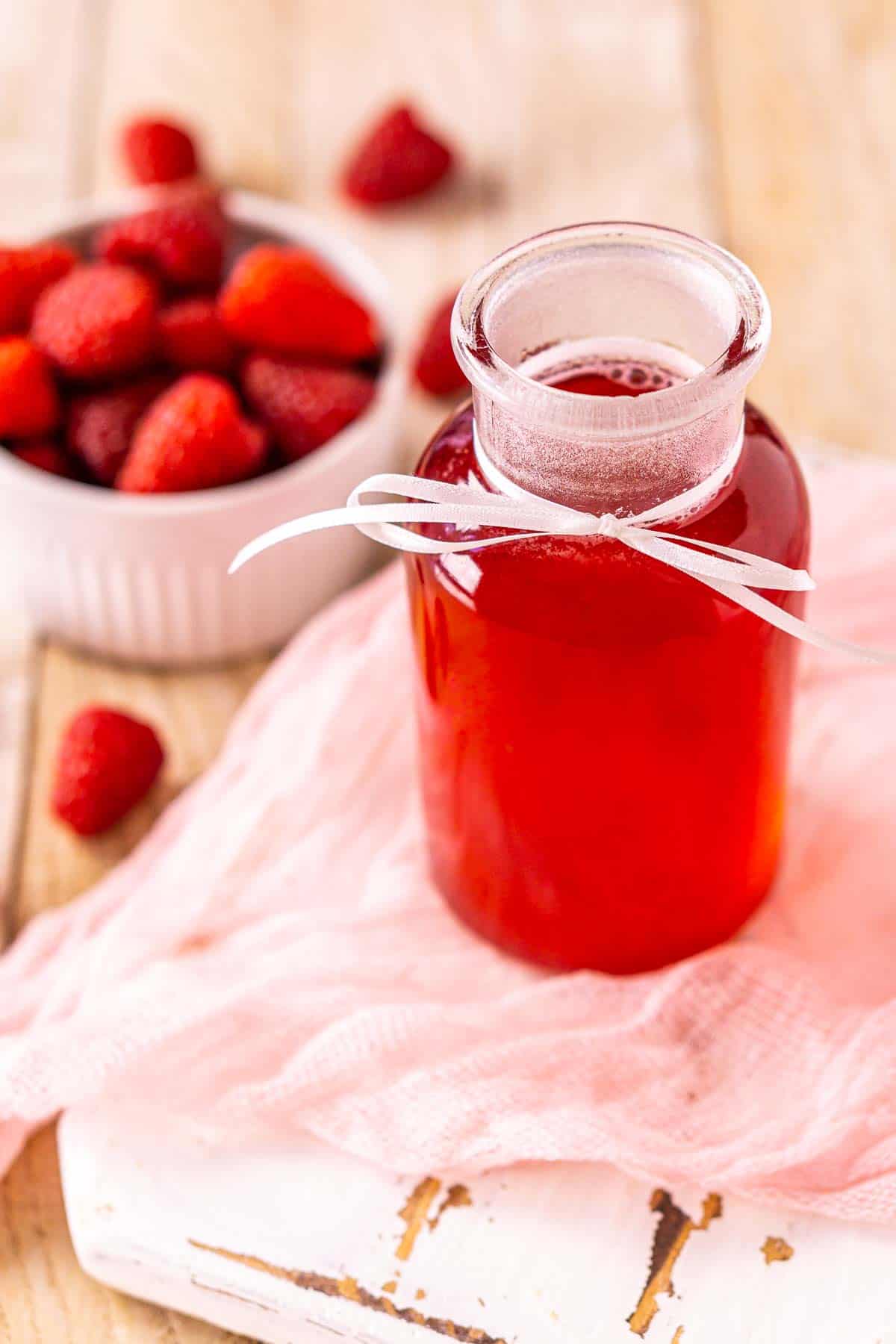 A bottle of raspberry simple syrup on a white tray with a piece of pink clothe and a small bowl of berries to the left.
