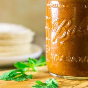 A close-up of a mason jar filled with the red enchilada sauce on a wooden tray with cilantro leaves around it.