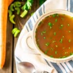 A bowl of seafood stock on a white and blue napkin with a carrot, parsley and spoon to the side on a wooden surface.