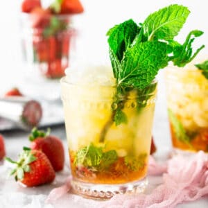 The strawberry mint julep on a pink clothe with a cup full of strawberries in the background.