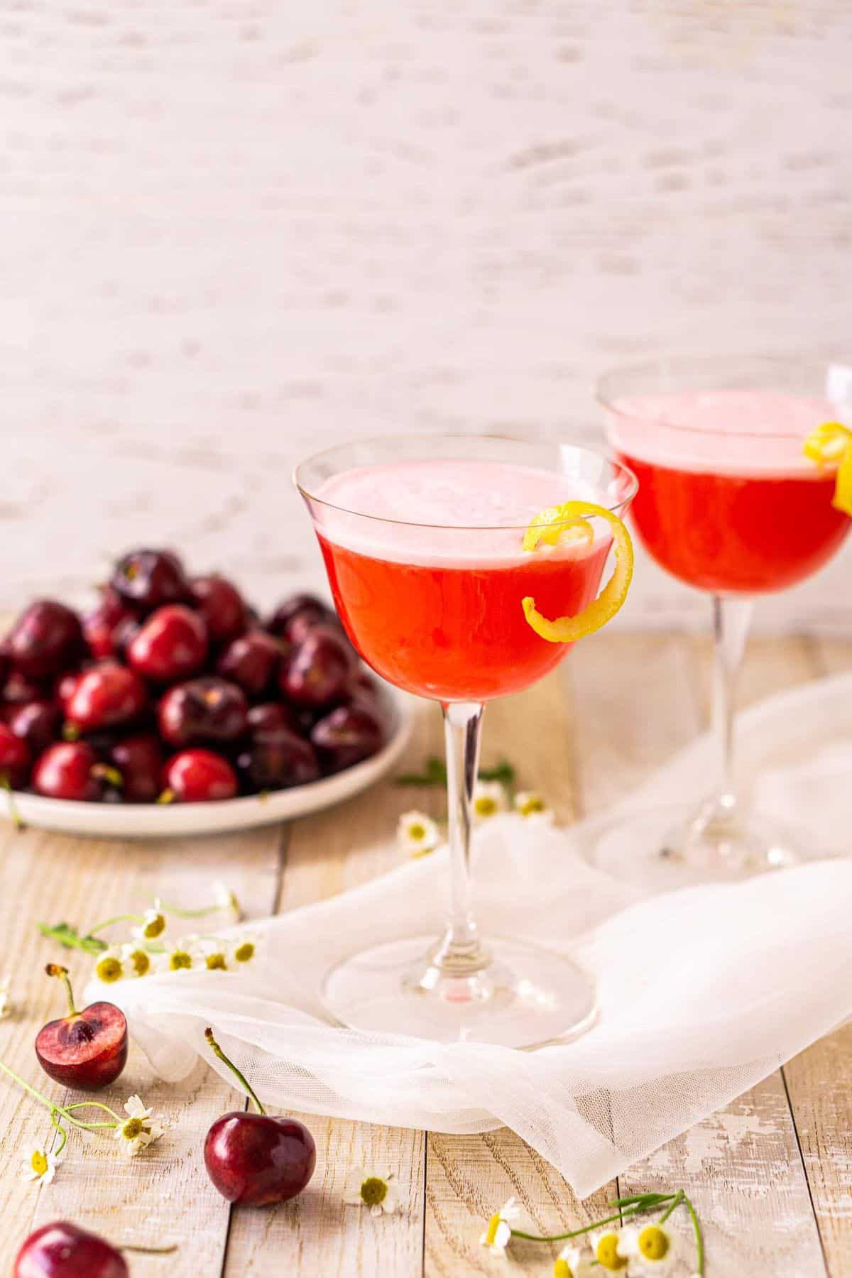 Two cherry vodka sours on a cream-colored wooden surface with cherries and white flowers around them.