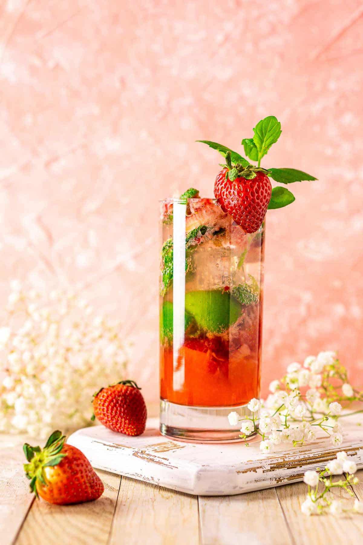 A strawberry mojito on a white wooden serving tray with strawberries and flowers around it against a pink background.