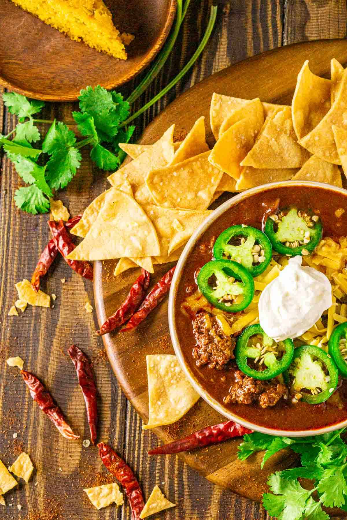 An aerial view of a bowl of venison chili with a slice of cornbread, dried chiles, cilantro and tortilla chips to the side.