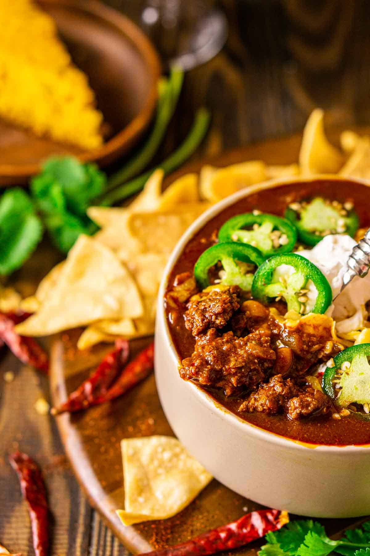 A spoon in the bowl of venison chili bringing the meat up with cilantro, chips and dried chiles scattered to the left.