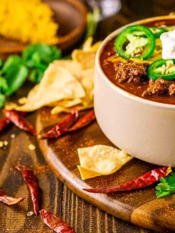A cream-colored bowl full of the venison chili on a brown wooden plate with crushed tortilla chips and dried chiles surrounding it.