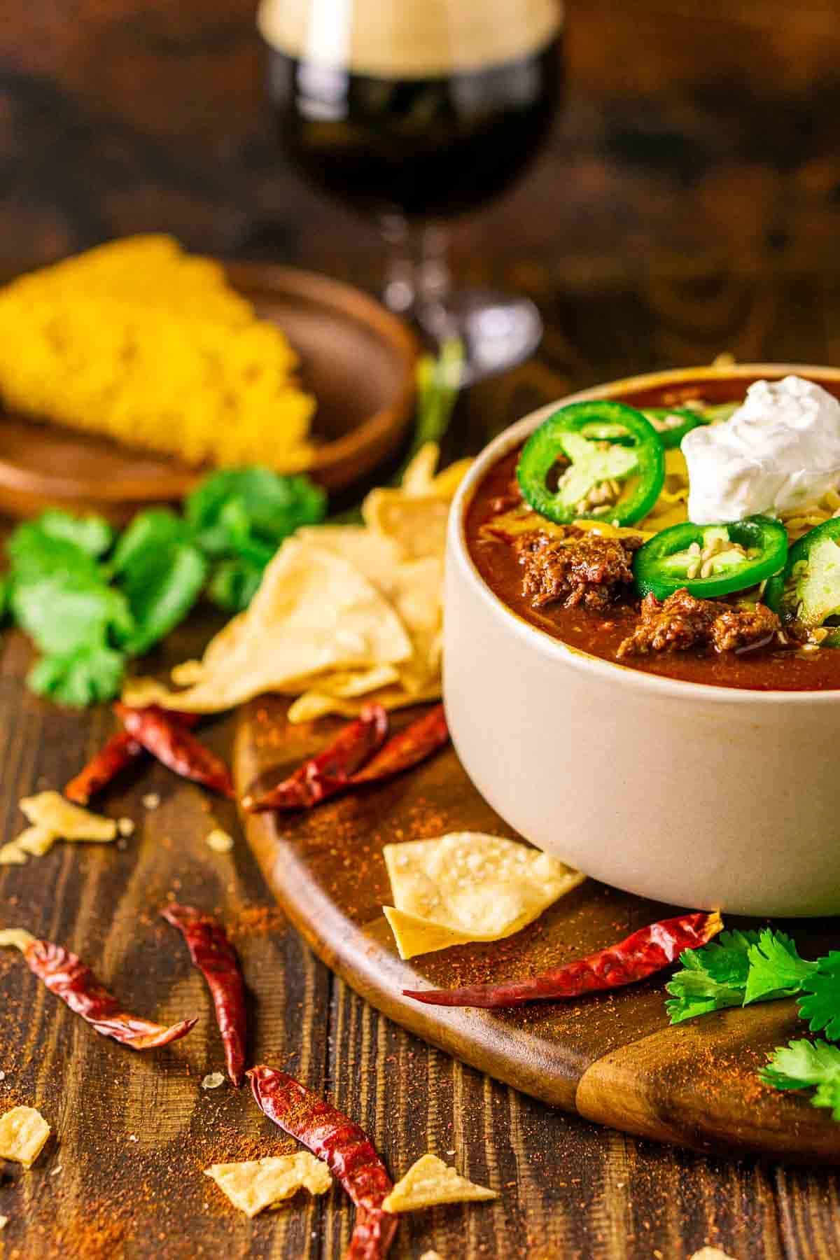 A cream-colored bowl full of the venison chili on a brown wooden plate with crushed tortilla chips and dried chiles surrounding it.