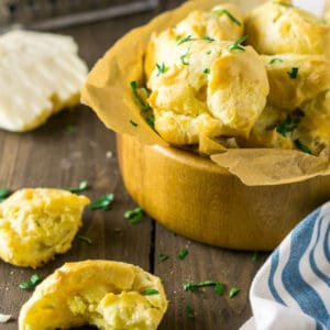 A wooden bowl filled with the white cheddar gougeres with grated cheese and chopped parsley scattered around it.
