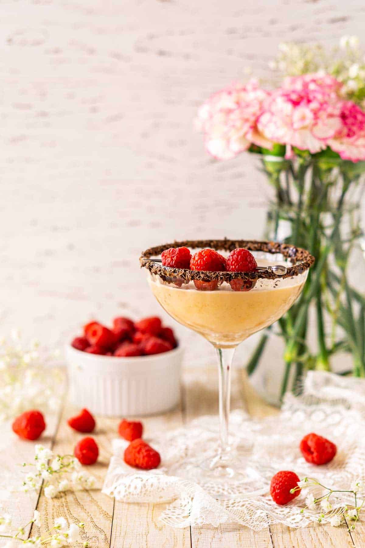 A straight-on shot of the chocolate-raspberry martini on white lace with raspberries and white flowers scattered around it.