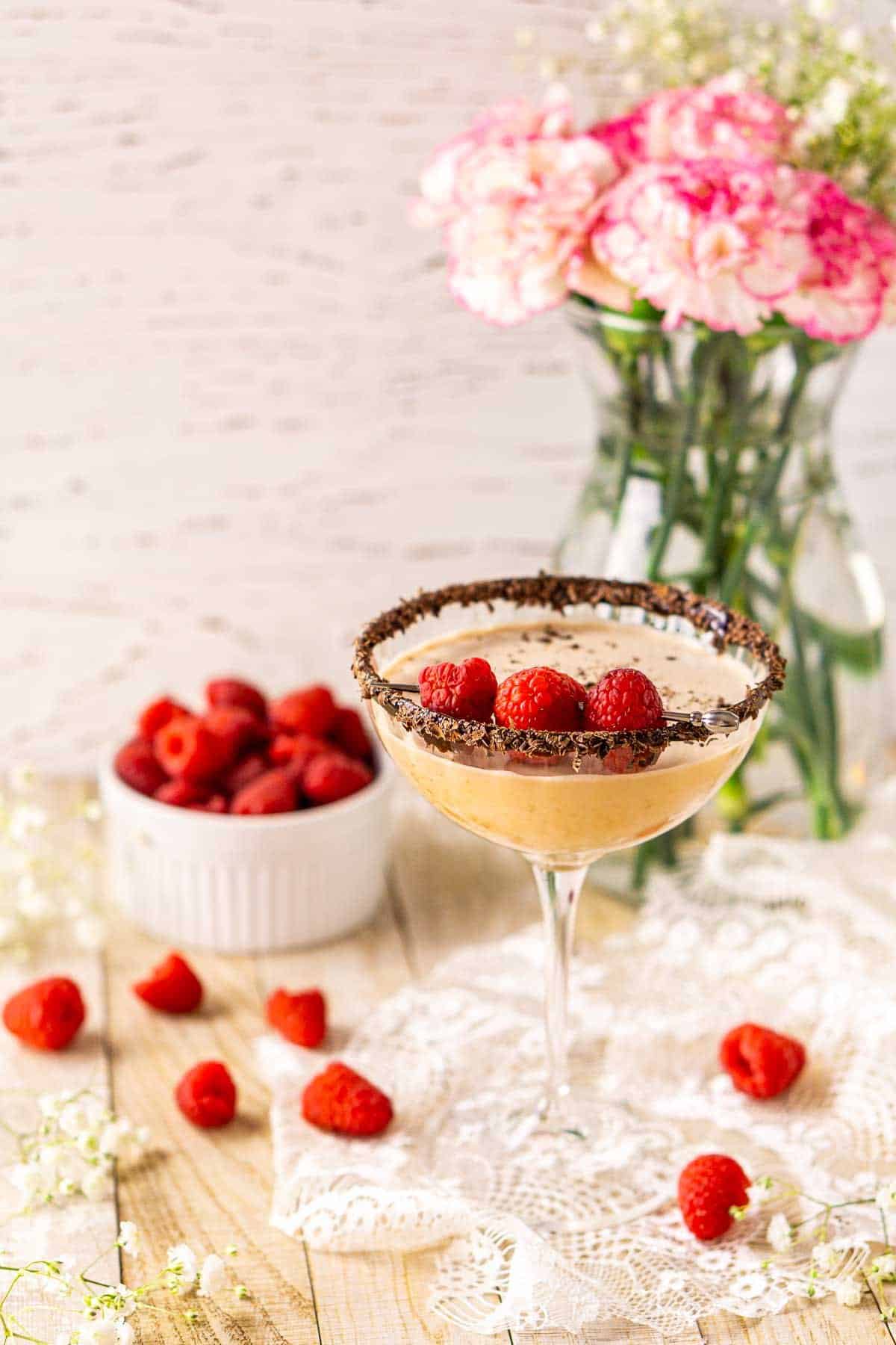 Looking down on a chocolate-raspberry martini on a piece of white lace with a bowl of raspberries and a vase of flowers behind it.