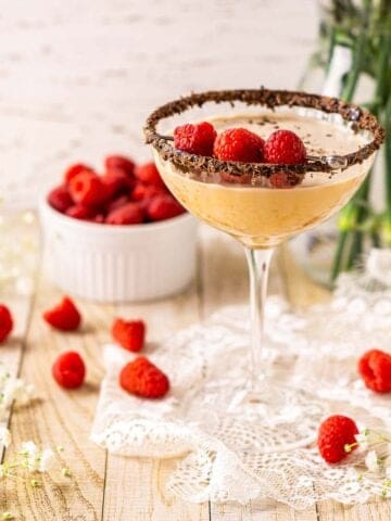 A chocolate-raspberry martini on a piece of white lace with raspberries scattered around it and flowers in the background.