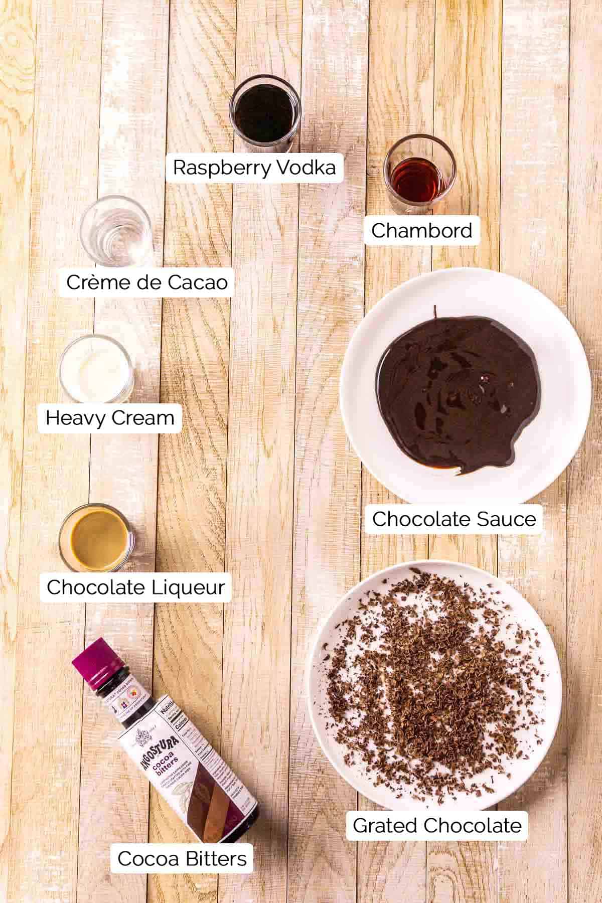 The martini ingredients with black and white labels by each item on top of a lightly colored wooden board.