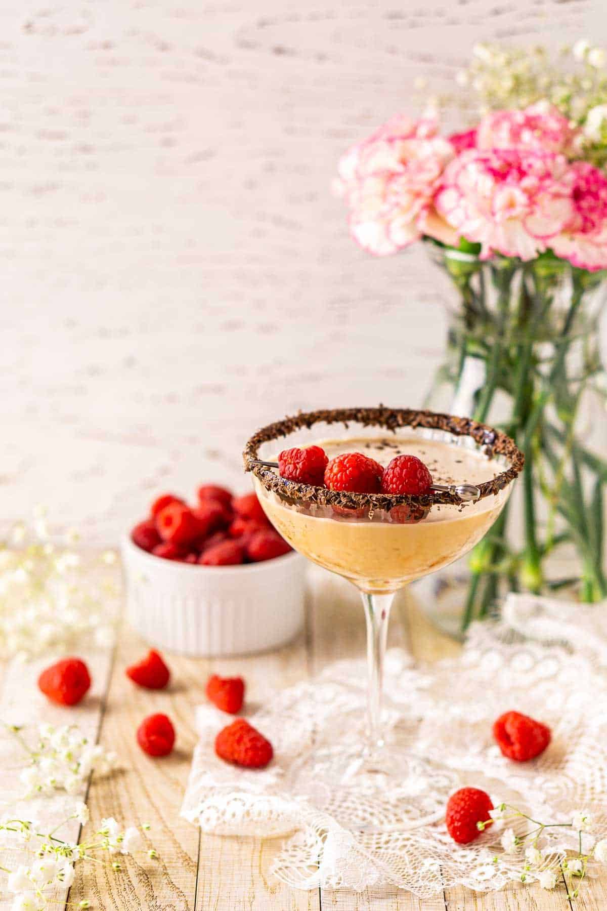 A chocolate-raspberry martini on a piece of white lace with raspberries scattered around it and flowers in the background.