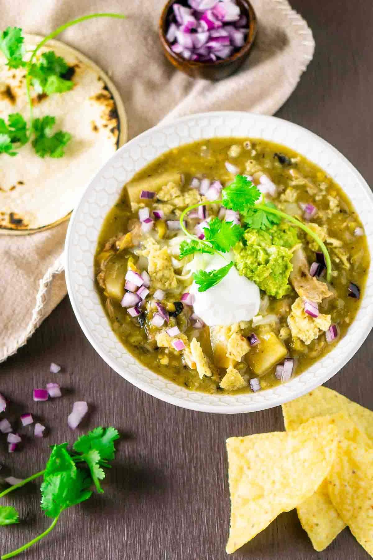 An aerial view of the Colorado pork green chili on a brown surface with tortillas, cilantro, red onion and chips scattered around it.