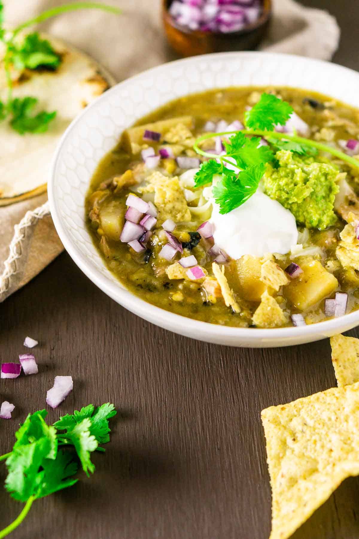 Looking down on a bowl of Colorado pork green chili with a stack of flour tortillas to the left and a small bowl of red onions in the background.