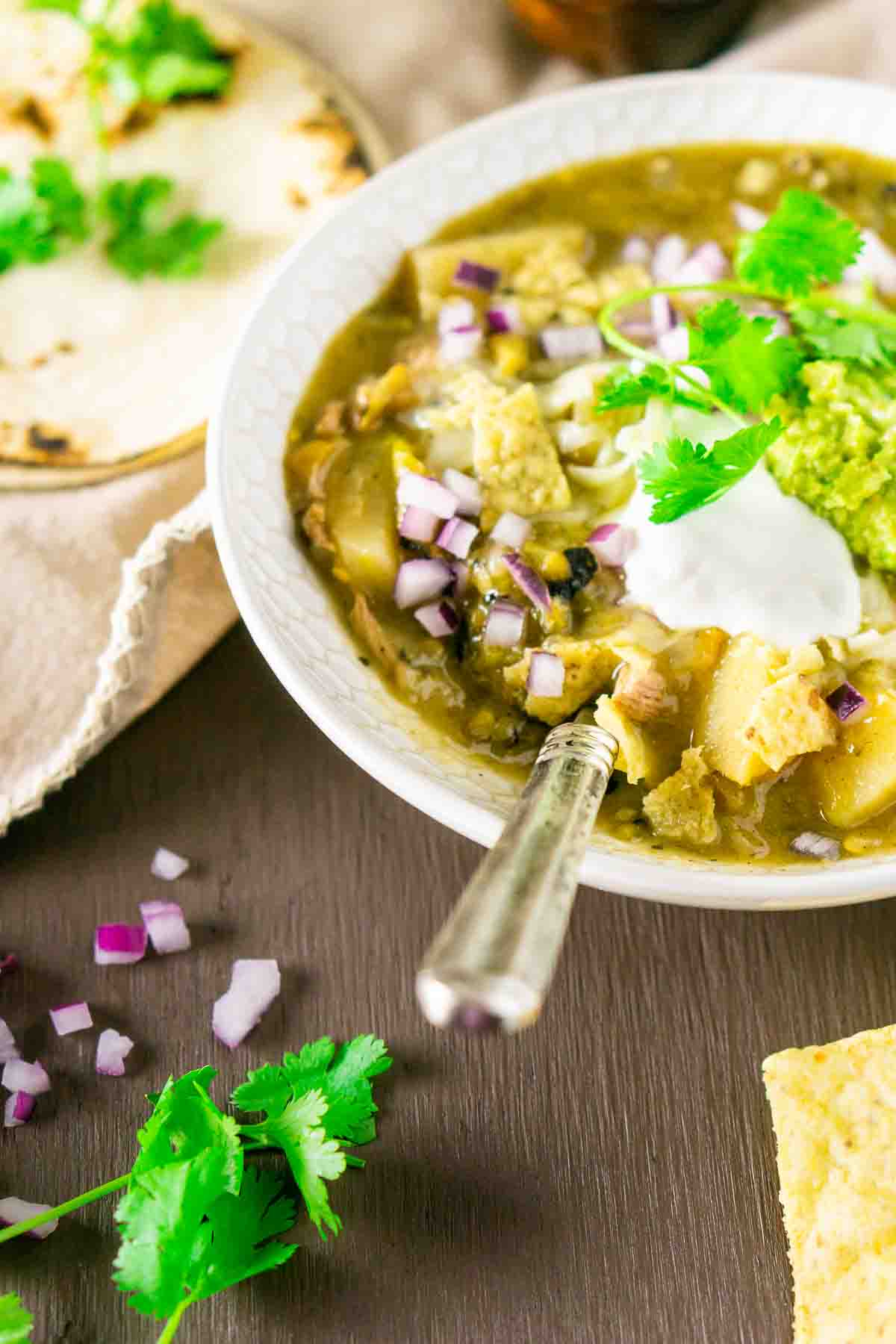 A bowl of Colorado pork green chili with a spoon sticking out and tortilla chips to the right and a stack of tortillas, chopped red onions and sprigs of cilantro to the left.