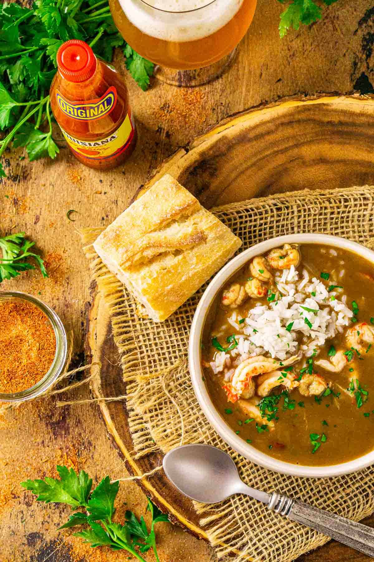 An aerial view of a bowl of crawfish gumbo with sprigs of parsley, a bottle of hot sauce and a beer to the left.