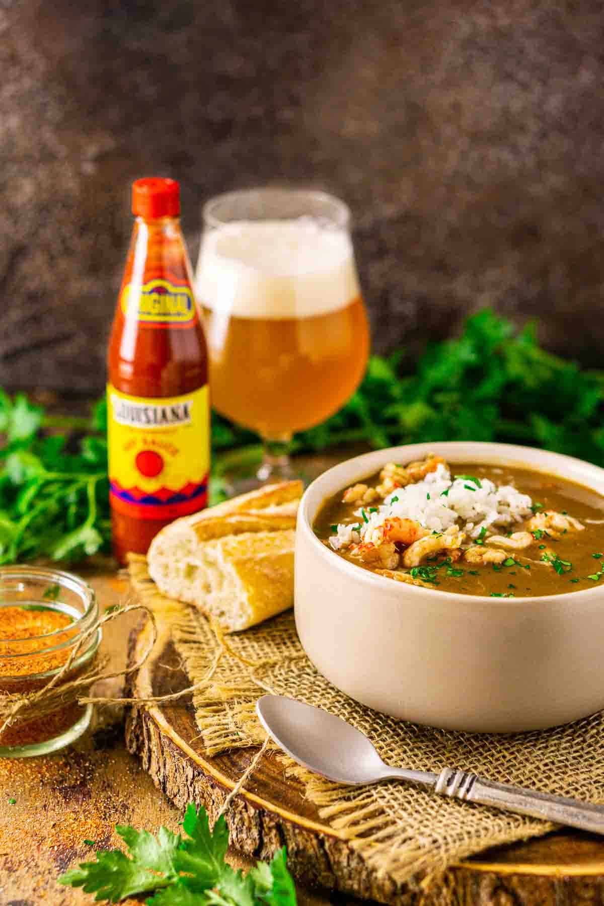 A bowl of the crawfish gumbo on a wooden platter with a spoon and burlap and a jar of Cajun seasoning to the left.