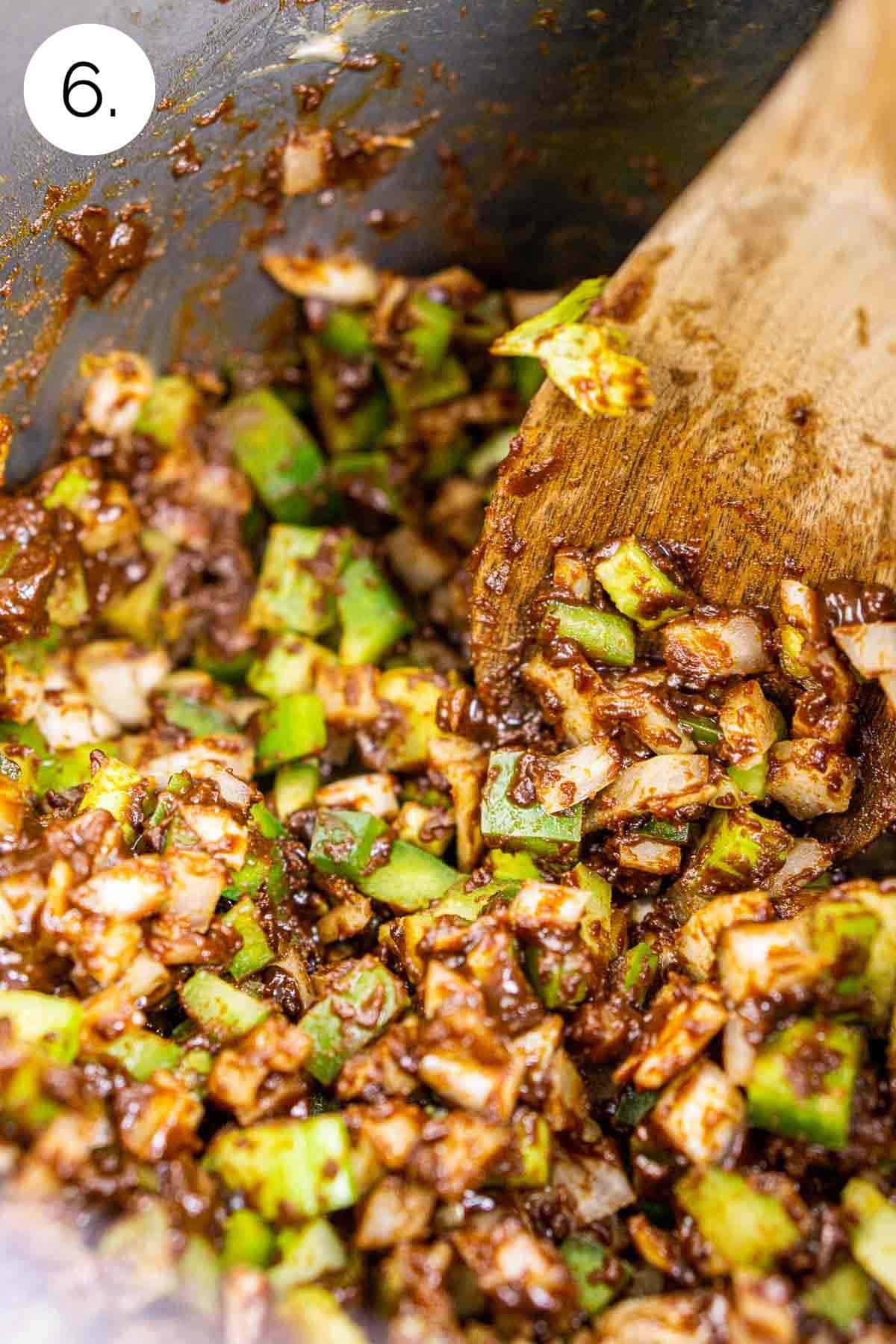 A wooden spoon stirring the green bell pepper, onion and celery with the dark brown roux in a large stainless steel stock pot.