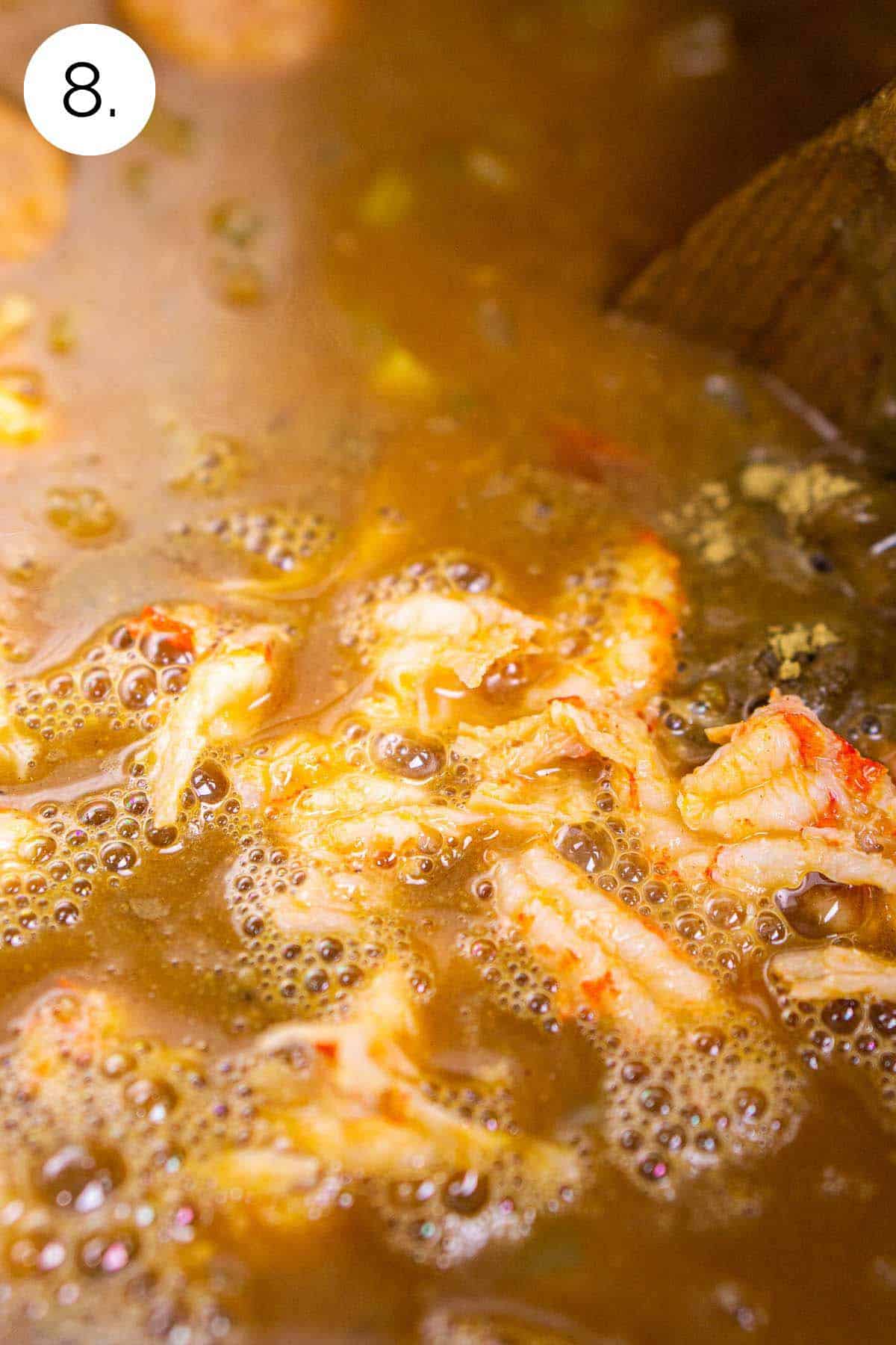 A wooden spoon stirring the crawfish into the broth on the stove-top range in a large stock pot.