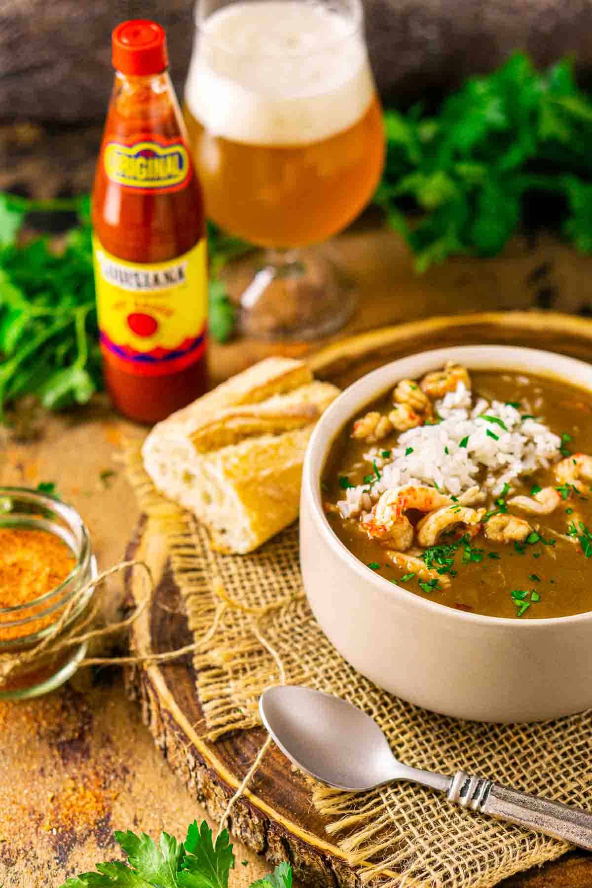 Looking down on a bowl of crawfish gumbo with a silver spoon in front and a bottle of hot sauce and beer in the background.