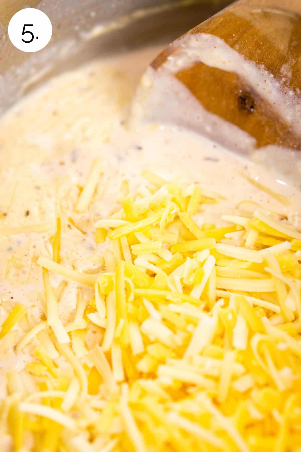 A wooden spoon stirring the shredded cheese into the cream mixture to make the sauce.