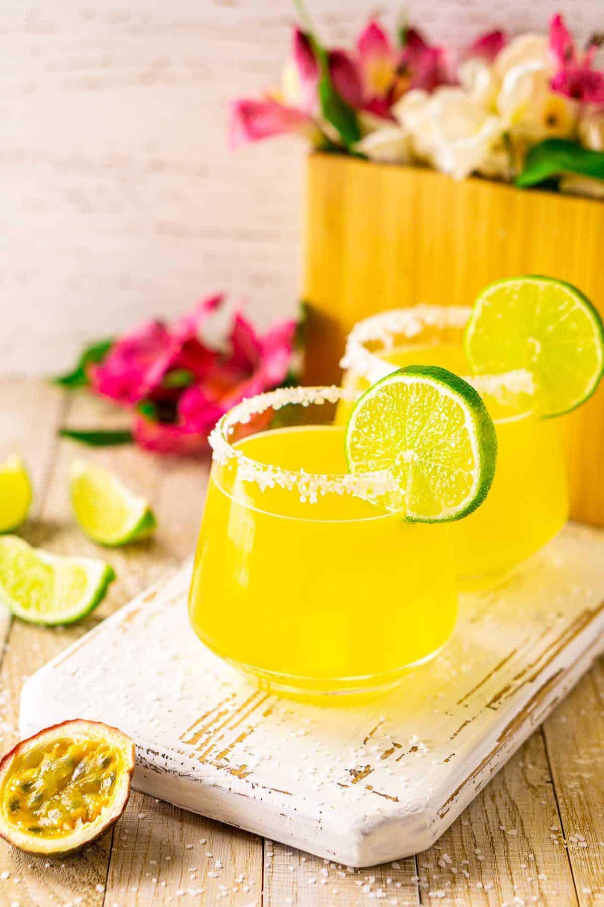 An aerial view of a couple passion fruit margaritas on a white serving tray with tropical flowers in the background.