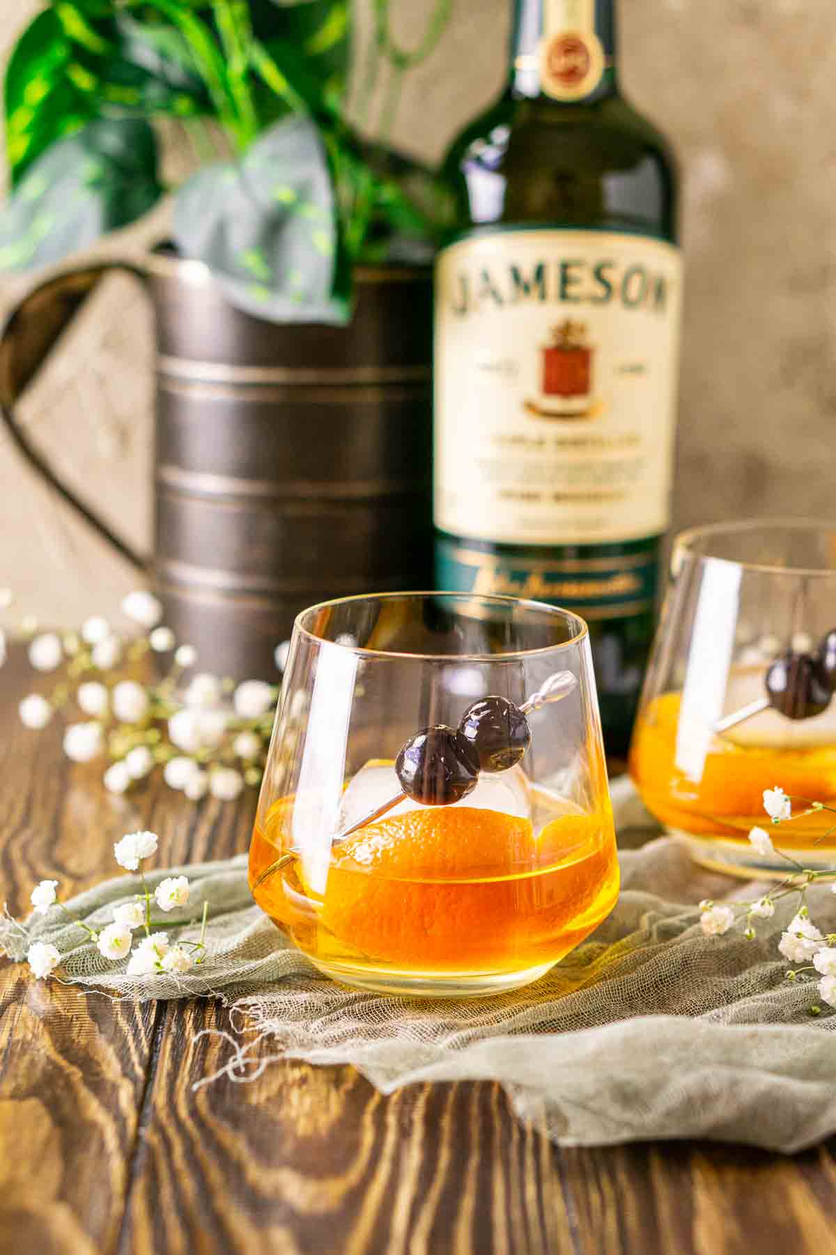 Looking straight on to an Irish old fashioned on a piece of green cloth with white flowers around it on a brown wooden board.