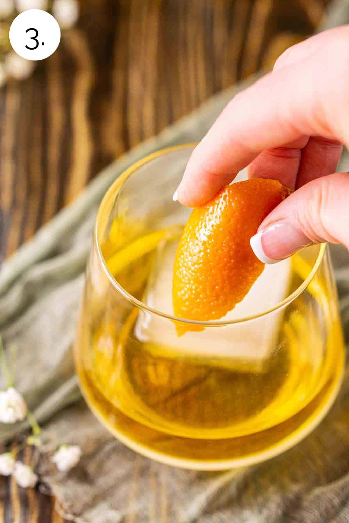 A hand adding an orange twist to the old fashioned glass to garnish after stirring and before serving.