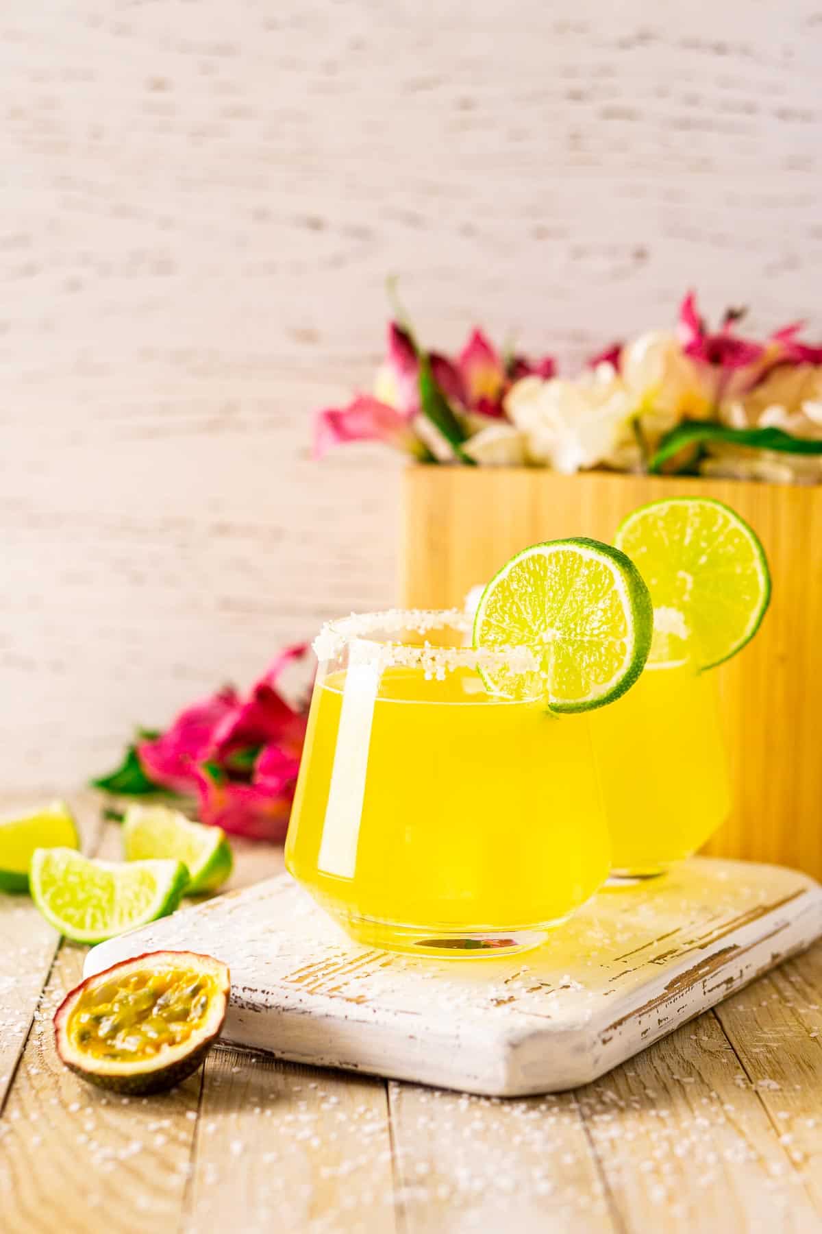 Two passion fruit margaritas on a small white wooden tray with limes and a cut passion fruit on the left.