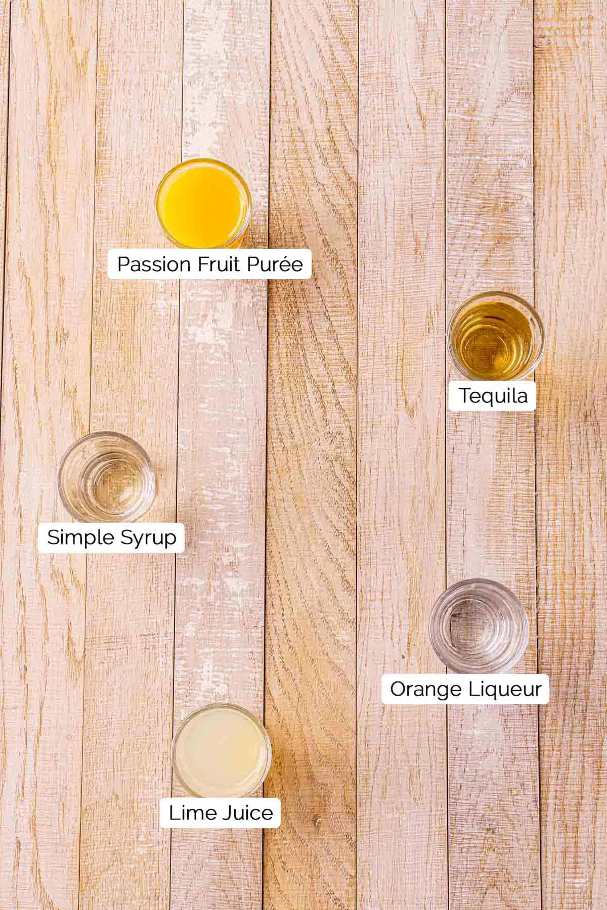 The drink ingredients in shot glasses on a cream-colored wooden board with black and white labels underneath each item.