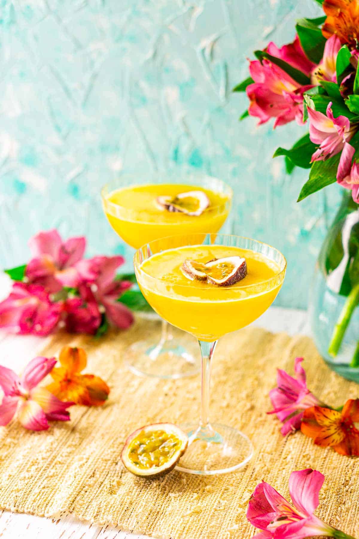 A side view of two passion fruit martini drinks on a straw placemat with a passion fruit and flowers to the side.