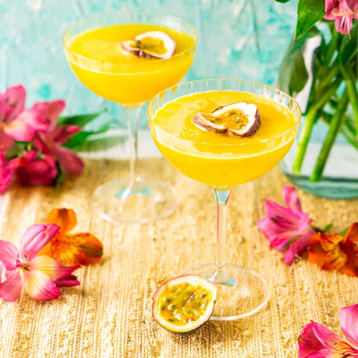 Two passion fruit martinis on a straw placemat with colorful flowers around them.
