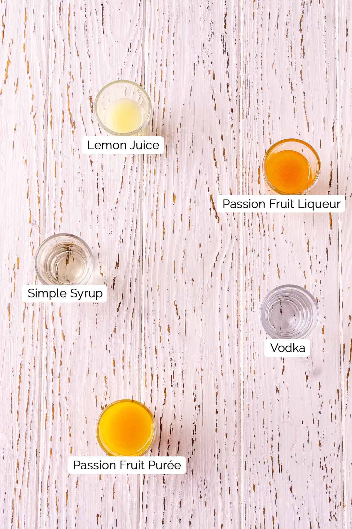 The ingredients in shot glasses on a white wooden board with black and white labels underneath each item.