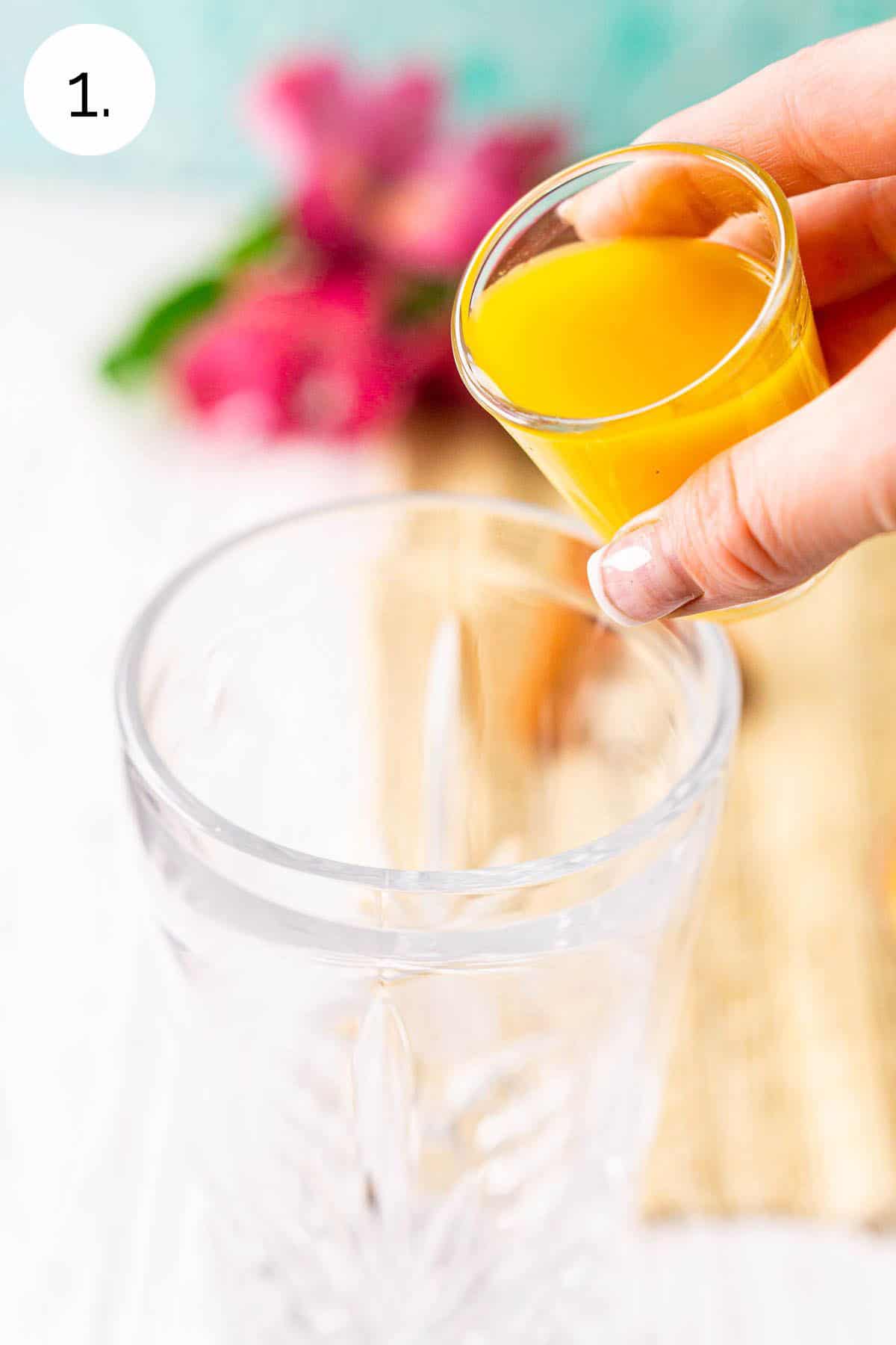 A hand pouring in a shot glass filled with passion fruit purée into a crystal cocktail shaker against a white background.