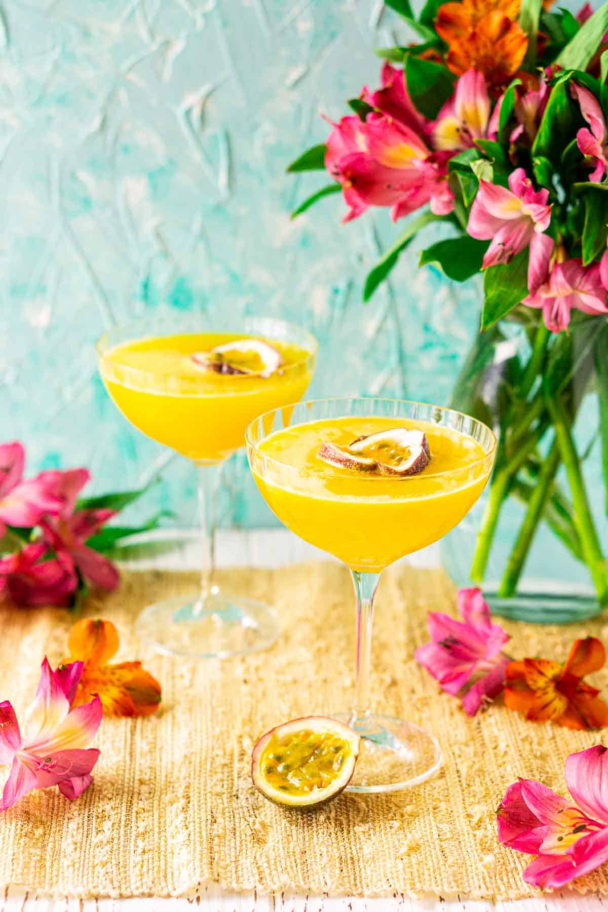 Two passion fruit martini cocktails on a straw placemat against a blue background with tropical flowers surrounding them on all sides.