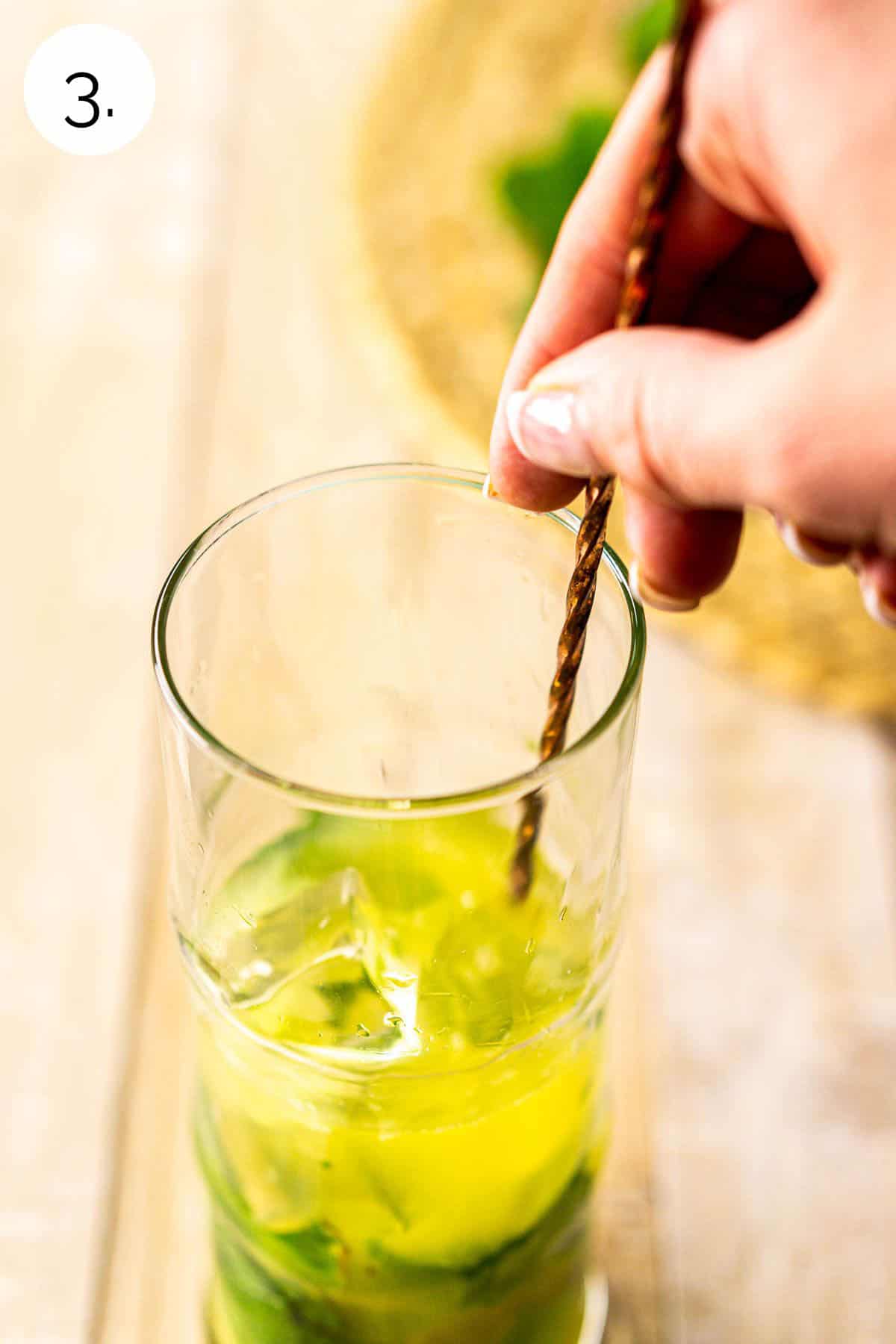 A hand stirring a copper bar spoon to mix in the white rum and ice in a tall highball glass on a cream-colored surface.