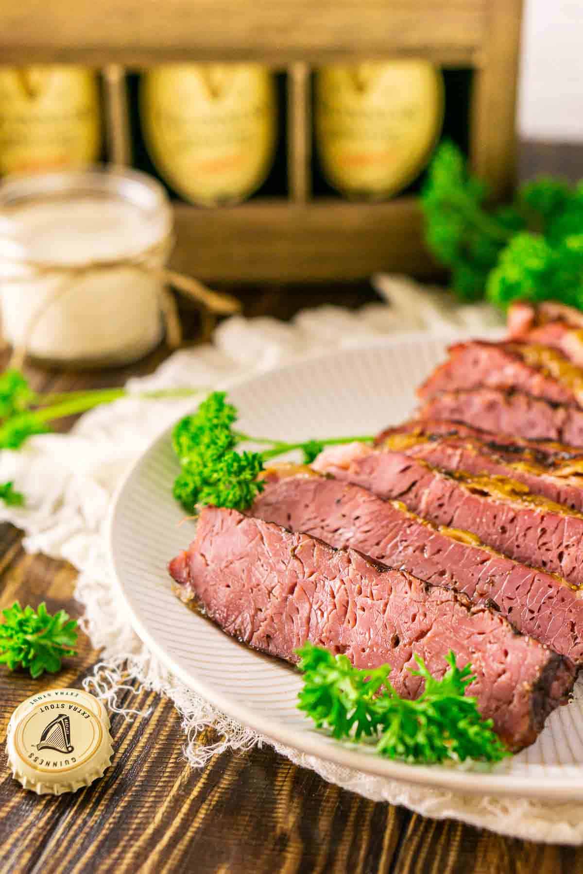 A side view of the smoked corned beef brisket on a white plate on top of a cream-colored clothe with fresh parsley sprinkled around it.