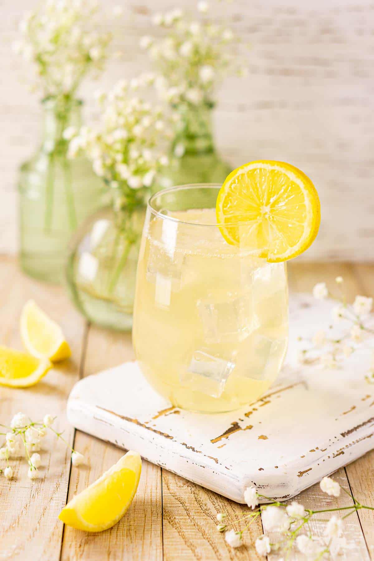 A glass of the St. Germain elderflower spritz on a wooden tray with white flowers and lemon slices in front.