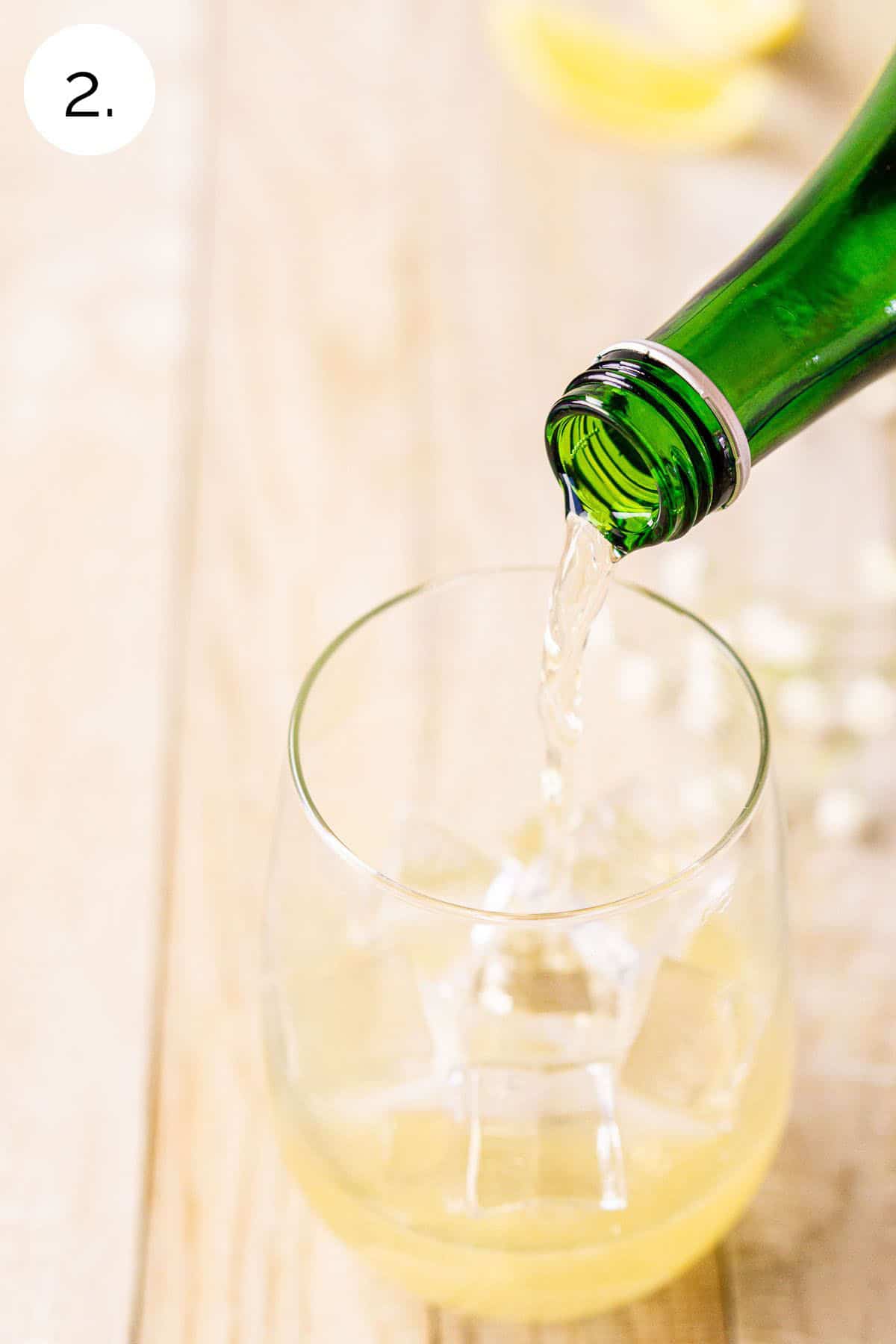 A Champagne bottle pouring white wine into the serving glass with a handful of ice on a cream-colored board.