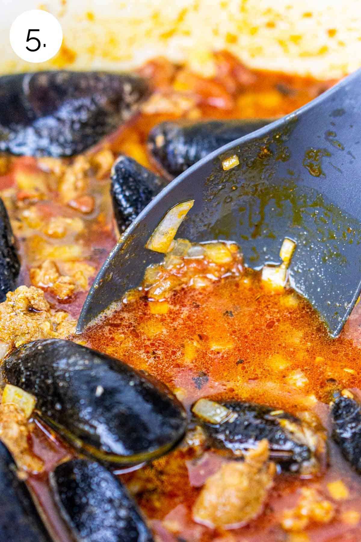 A black spoon stirring the mussels in a large Dutch oven after they've been added to the beer and tomato mixture.