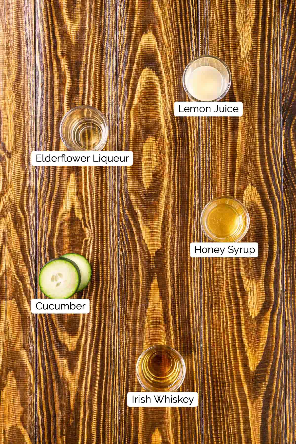 The drink ingredients in shot glasses on a brown wooden surface with white and black labels underneath the five items.