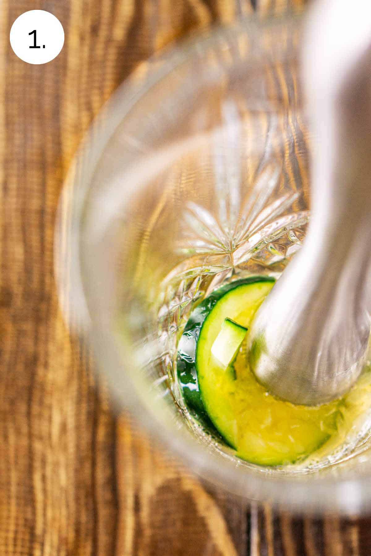 A stainless steel muddler breaking down the cucumber slices with the honey syrup in a clear cocktail shaker on a wooden surface.